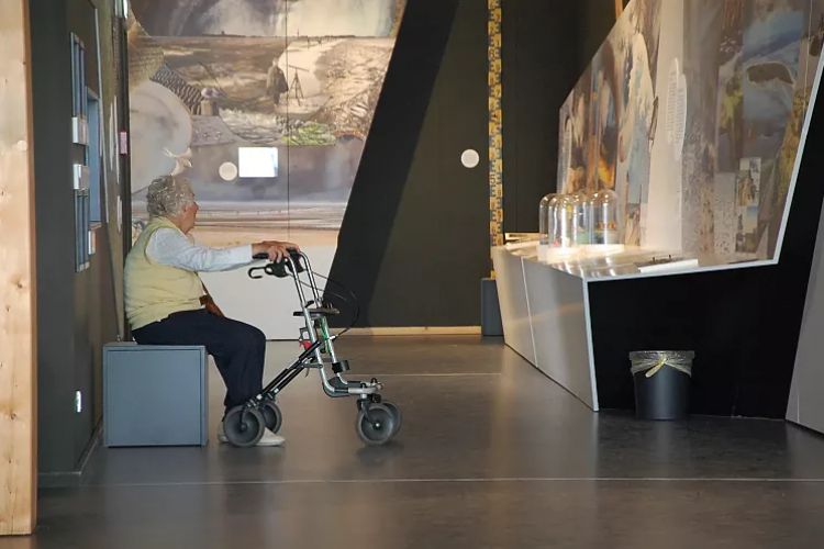 Eine alte Dame mit Rollator sitzt im Museum vor einem  Ausstellungsstück