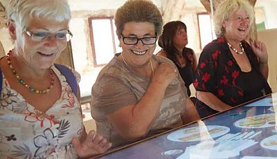 Drei ältere Damen sitzen lächelnd vor einem multimedialen Tisch im Museum