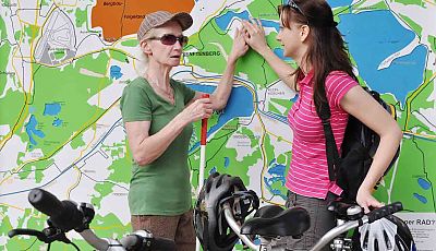Zwei Touristinnen mit Tandem, eine davon mit Blindenstock, ertasten eine taktile Karte der Seenlandschaft